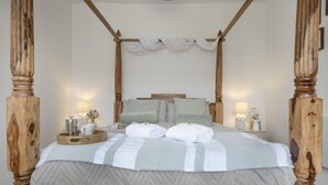 Una cama con dosel de madera, un dosel blanco y una colcha de color claro. Hay dos mesitas de noche con lámparas y objetos decorativos.