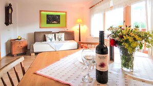 Een hotelkamer met een bed, nachtkastje, lamp en een eettafel met een wijnfles en bloemen.