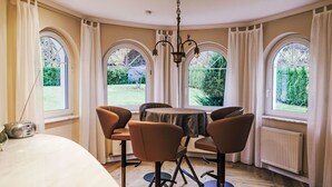 Un comedor con una mesa redonda y cuatro sillas, ventanas arqueadas amplias, una araña y un radiador.