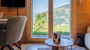 A room with a wooden interior, a television mounted on the wall, a table with a remote control, and a view of a mountainous landscape through a large sliding glass door. - Cosy apartment for 4 guests with WIFI, TV and terrace (Basse-nendaz)