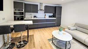A modern kitchen with dark cabinetry, a white countertop, and a built-in oven. There is a dining area with a table and chairs, and a sofa in the living area. - Beautiful apartment for 4 people with WIFI and TV (Nairn)