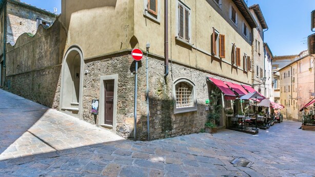 A narrow cobblestone street with a building featuring a stone facade, a red and white sign, and outdoor seating with umbrellas. - Beautiful apartment for 7 people with WIFI and patio (Volterra)
