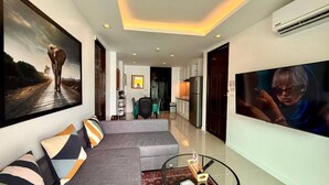 Una sala de estar moderna con un sofá, una mesa de centro y un televisor. Hay una zona de cocina con armarios y un refrigerador.