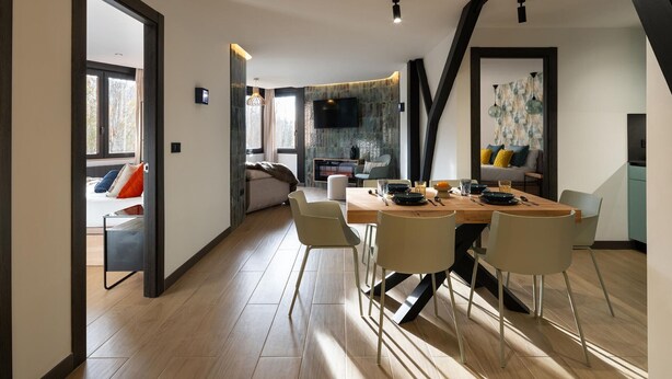A modern dining area with a wooden table and chairs, a kitchen in the background, and a cozy seating area with a sofa and cushions.