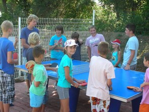 Eine Gruppe Menschen spielt draußen Tischtennis an einem blauen Tisch, im Hintergrund ist ein weißer Zaun zu sehen.