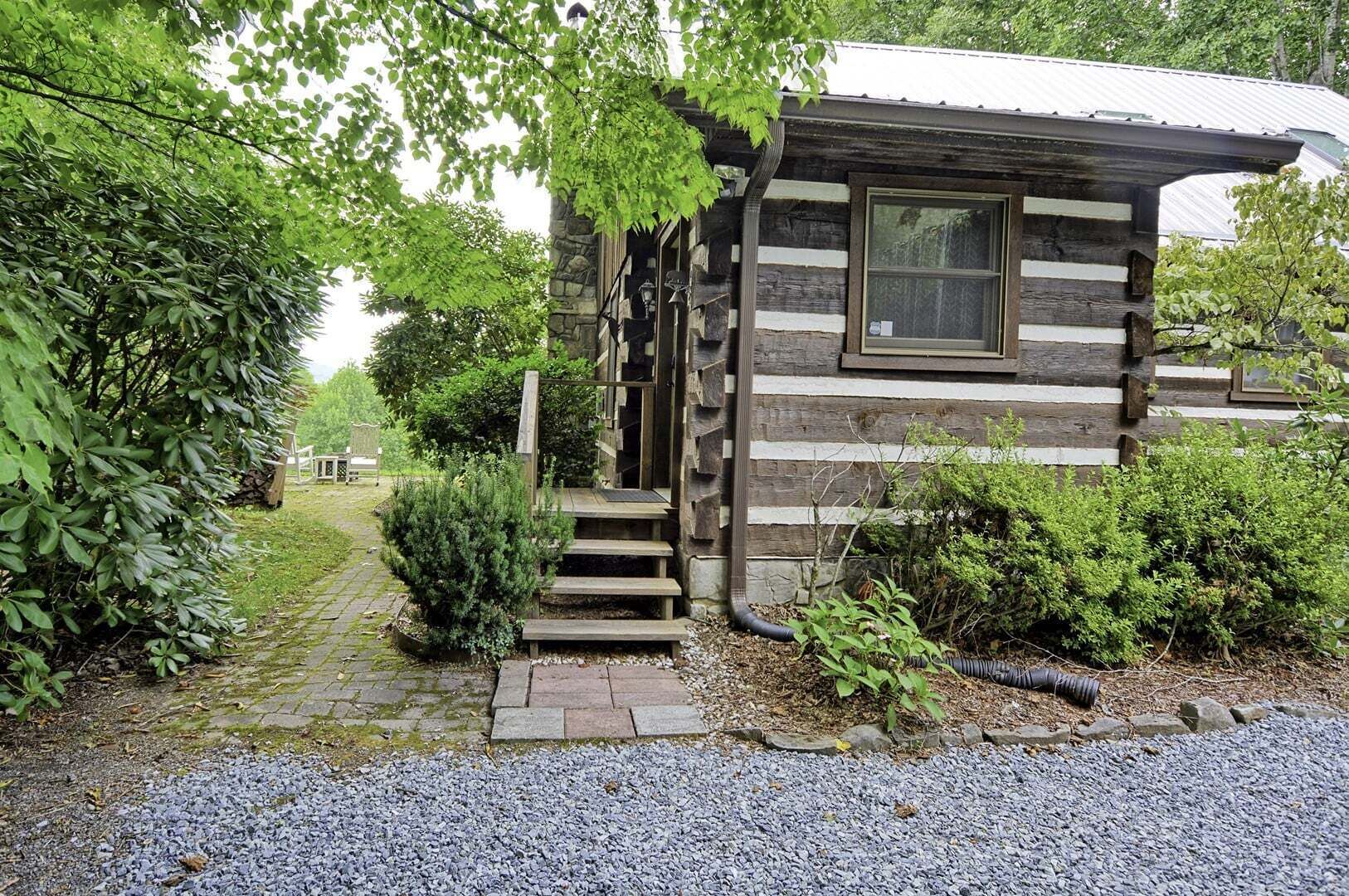 A rustic cabin with a wooden staircase leading to a door, surrounded by greenery and a gravel path.