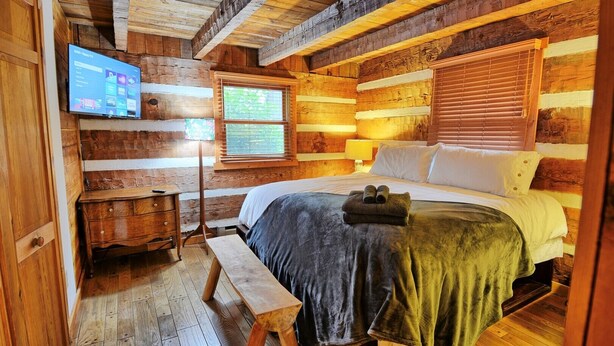 A cozy log cabin room with a bed, wooden walls, a desk, and a TV mounted on the wall.