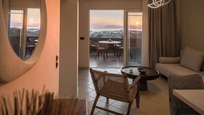 Una sala de estar moderna con un sofá, una mesa redonda para café y sillas de mimbre. Un espejo grande está en la pared, y se ve un atardecer a través de puertas de vidrio.