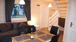 Una sala de estar acogedora con un sofá, una mesa de comedor con individuales, copas de vino y una botella. Se ve una ventana con cortinas, y una escalera conduce a otra habitación.