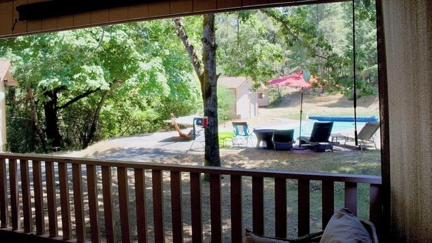 Blick von einer überdachten Veranda mit Holzbalkon auf einen Garten mit Schwimmbecken, Liegestühlen und einem roten Sonnenschirm.