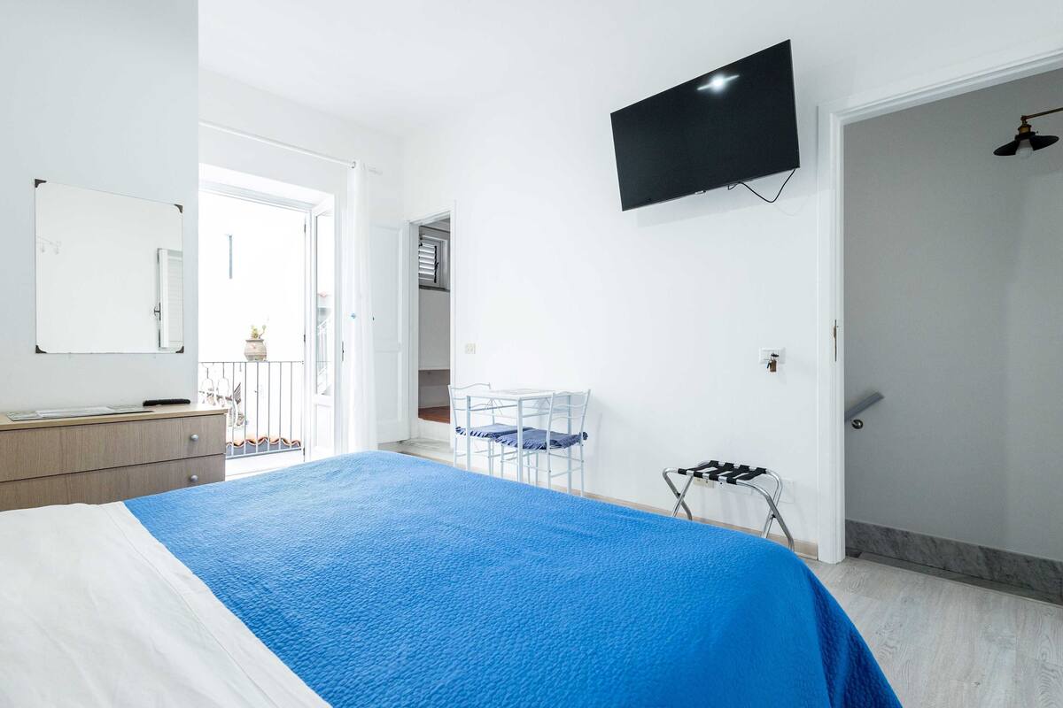A bedroom with a bed, a desk, a chair, a TV, and a door leading to a balcony.
