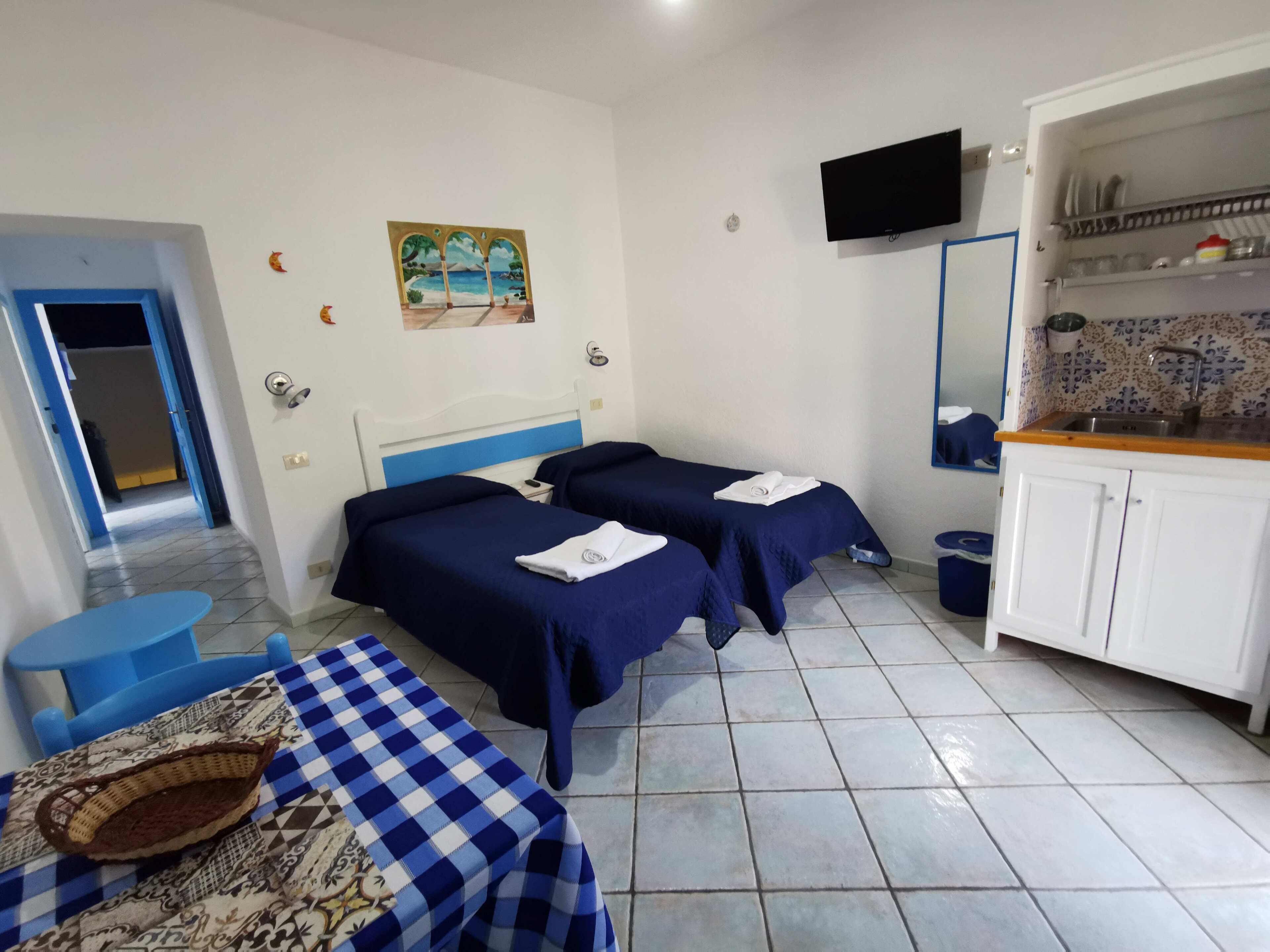 A hotel room with two beds, a small table, a TV, and a kitchen area.