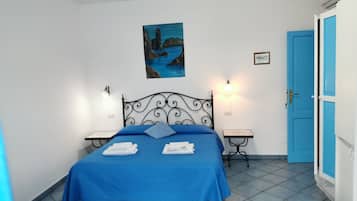 A bedroom with a bed, two bedside tables, a wall-mounted light, a painting, and a blue door.