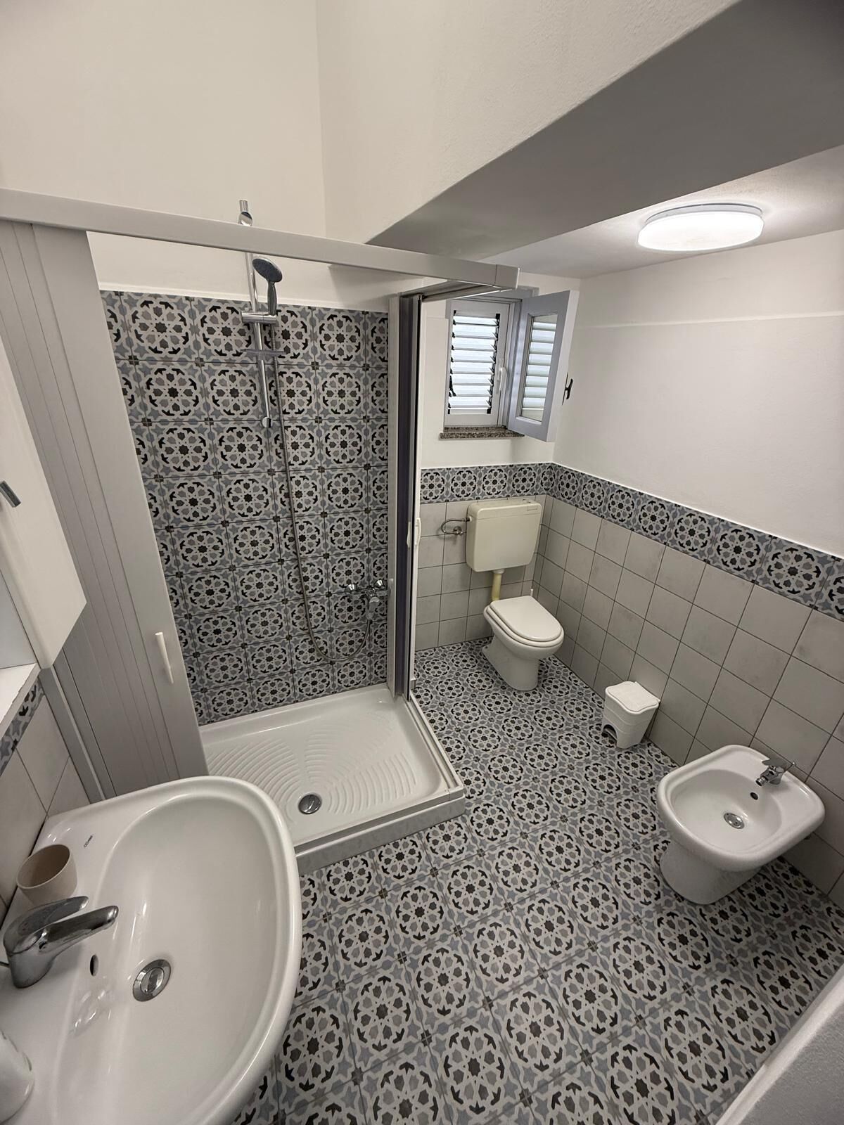 A bathroom with a shower, toilet, bidet, and sink, featuring patterned tiles and a small window.
