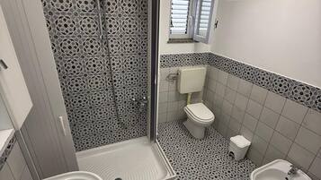 A bathroom with a shower, toilet, bidet, and sink, featuring patterned tiles and a small window.