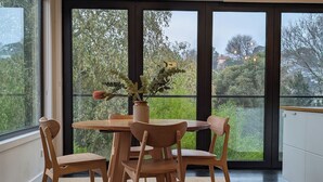 A dining area with a wooden table and chairs, a vase with flowers, and large windows offering a view of greenery and buildings. - Your CBD Treetop Base | Parking, Wifi & Views (Launceston City)