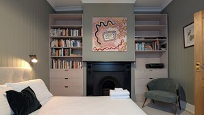A bedroom with a large bed, a fireplace, bookshelves, and a chair. - Your CBD Treetop Base | Parking, Wifi & Views (Launceston City)