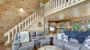 A spacious living area with a wooden staircase, a sofa set, and a kitchen in the background. - Stunning Mountain Cabin w/ Hot Tub & Decks (Blue Ridge)
