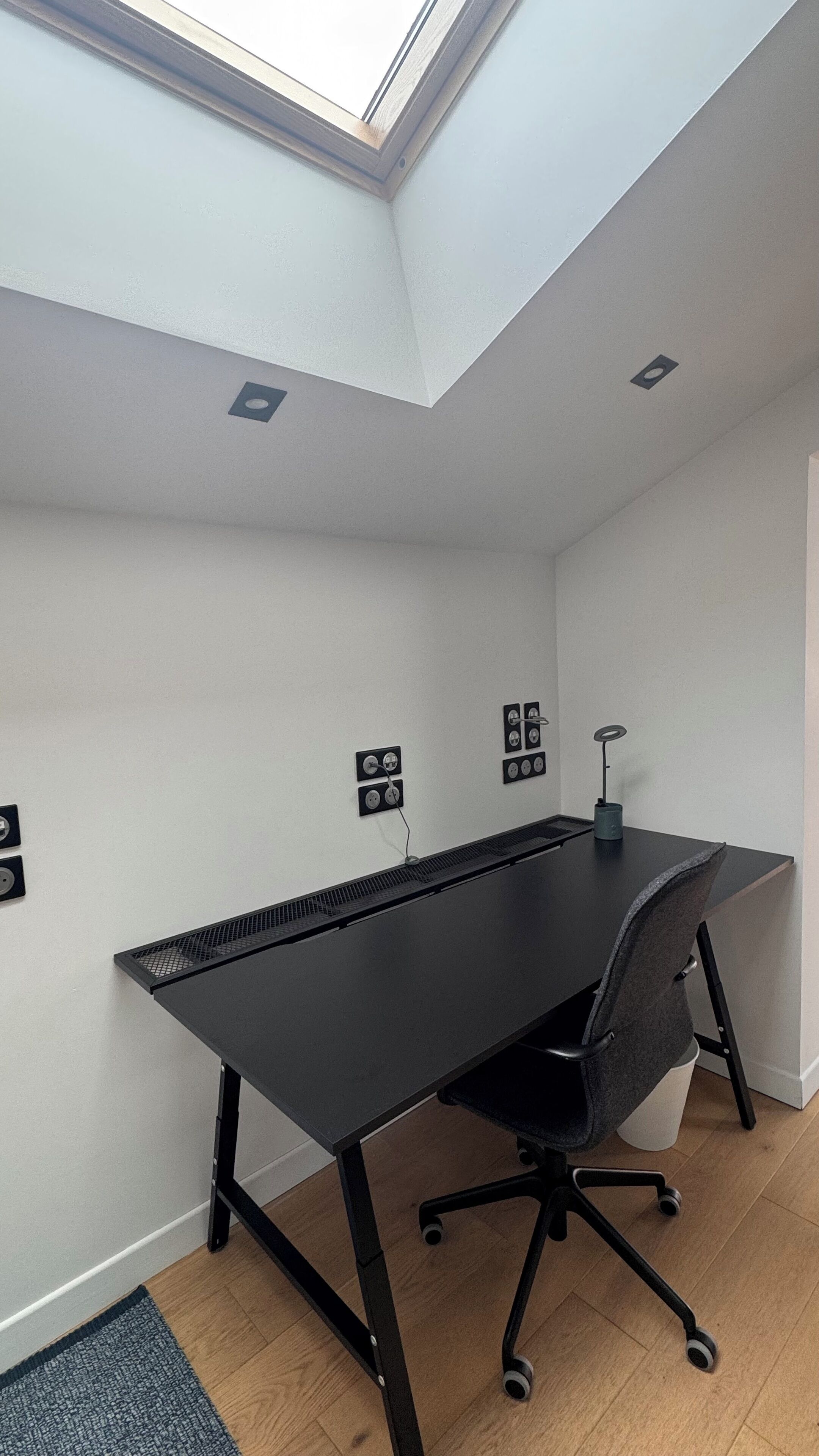 A modern office space with a black desk, a grey office chair, and a skylight.
