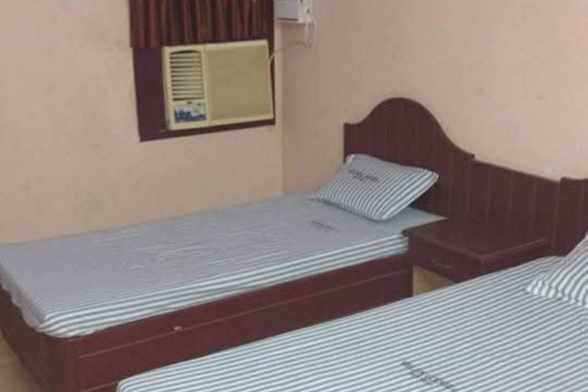 A room with two single beds, a small bedside table, a wall-mounted air conditioner, and a window with curtains.