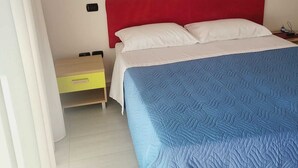 A bedroom with a red headboard, a blue quilted bedspread, a white pillow, and a yellow nightstand with a lamp. - By the sea in Miramare di Rimini (Rimini)