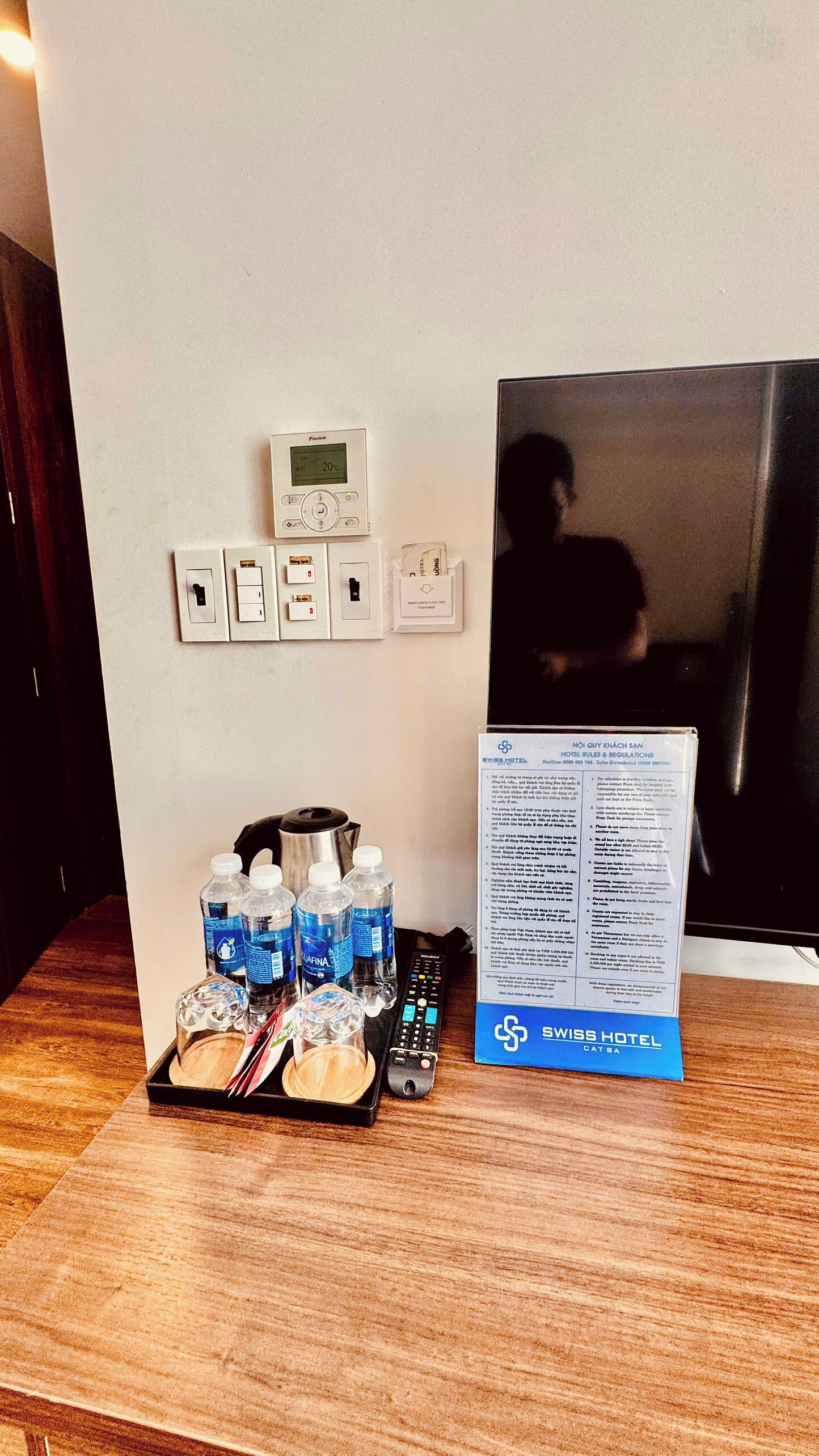 A hotel room with a thermostat, a power outlet, a water dispenser, and a remote control on a wooden table.