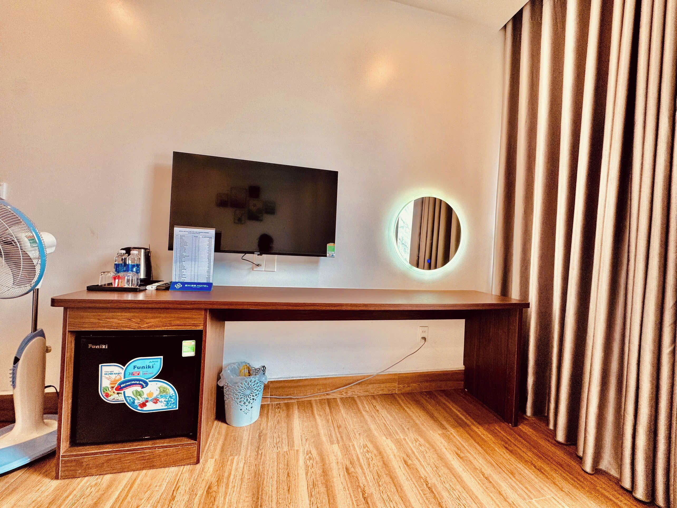 Habitación de hotel con un escritorio de madera, televisor, un pequeño cubo de basura y un ventilador de pie.