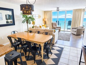 A dining area with a wooden table and chairs, a sofa, a lamp, and a view of the ocean through large windows.