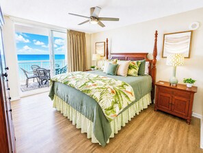 A bedroom with a bed, a nightstand, a lamp, a ceiling fan, a window with a view of the ocean, and a balcony with chairs.