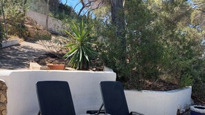 Deux fauteuils de jardin avec des structures métalliques et des assises en tissu maillé, placés sur une terrasse en pierre, avec un mur blanc et un petit arbre en arrière-plan.