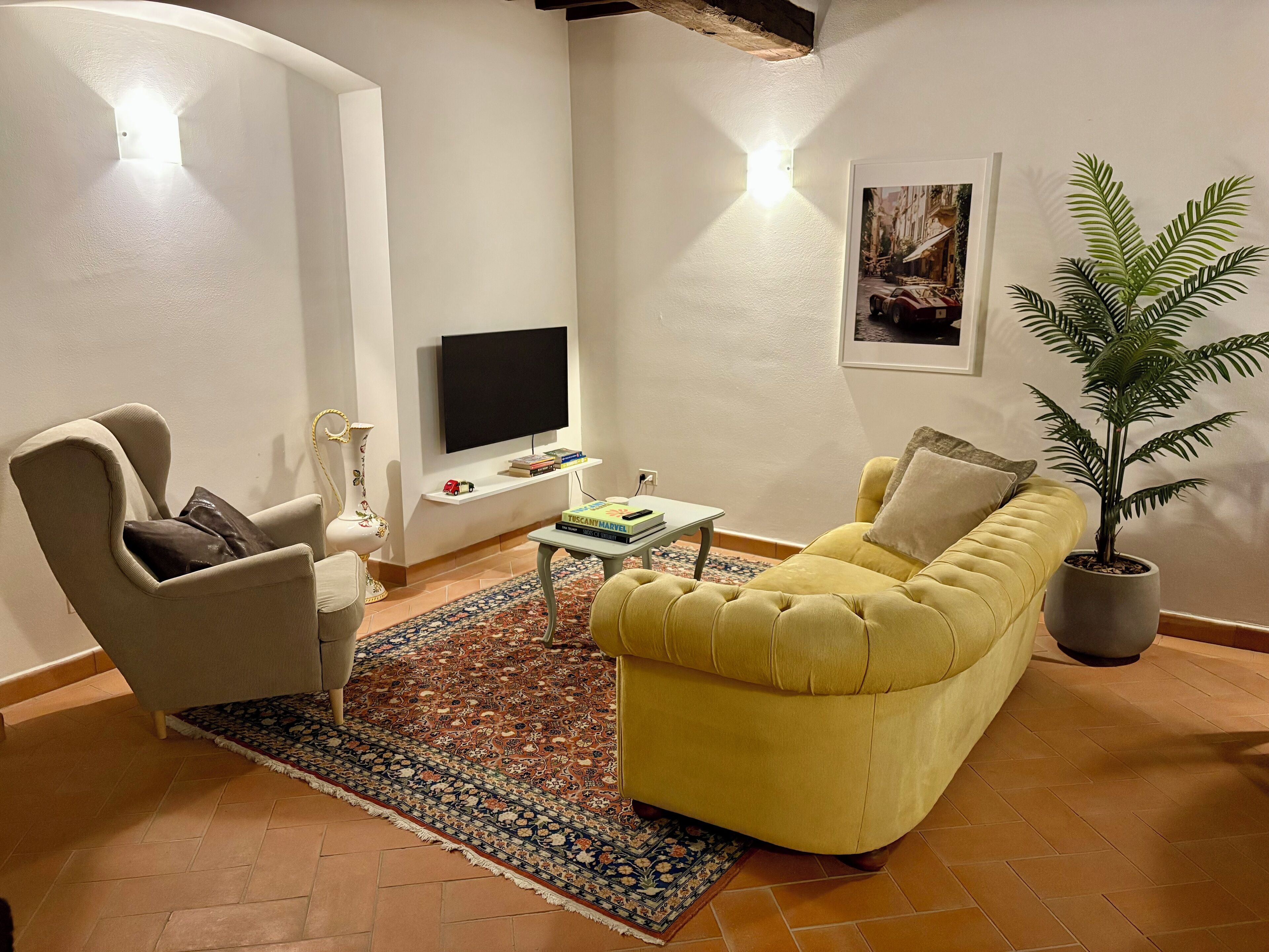 A living room with a yellow sofa, a white armchair, a small table with books, a TV on the wall, and a potted plant.
