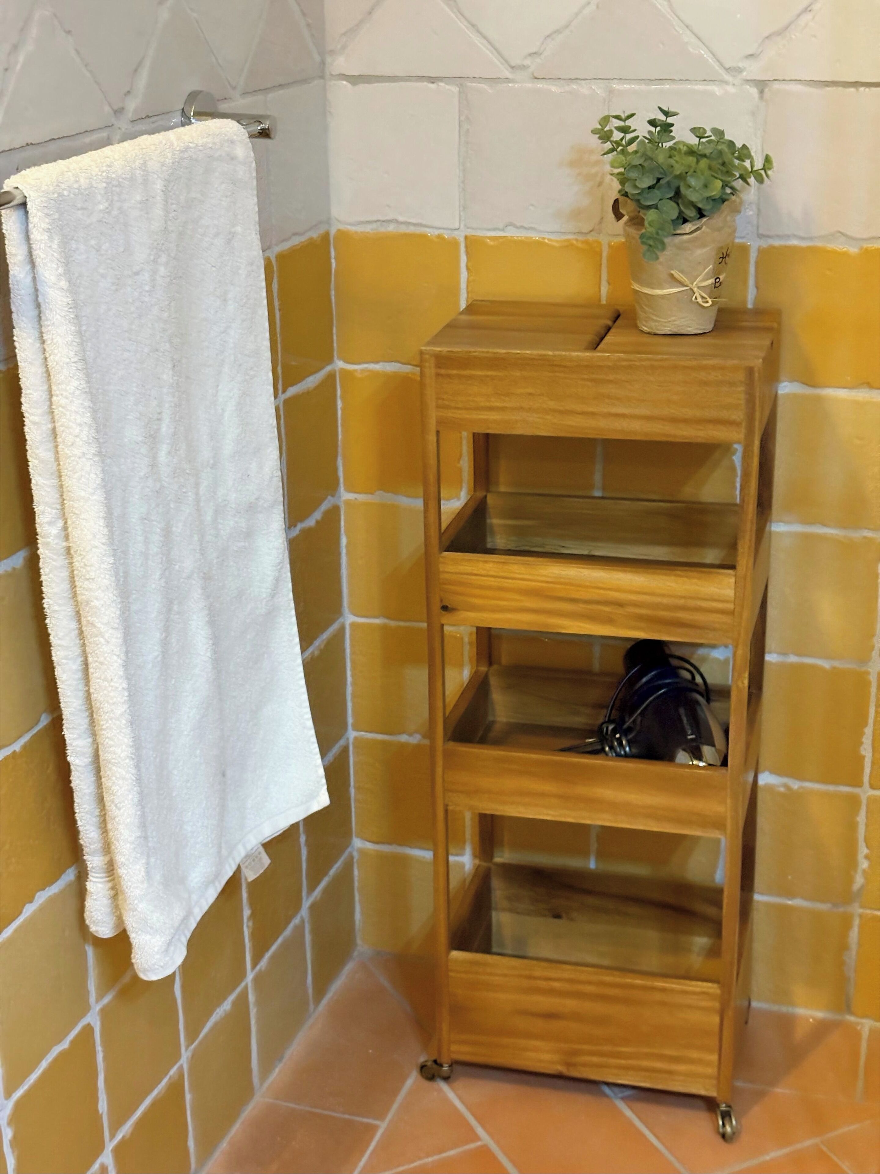 A bathroom with a white towel hanging on a rack, a wooden shelf unit with a potted plant on top, and yellow-tiled walls.