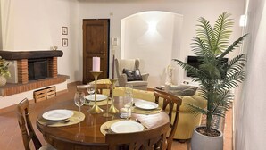 A dining area with a round wooden table set for four, surrounded by wooden chairs. A brick fireplace with a pink candle on a stand is visible in the background. - Condo Cortona, luxury apartment for 4 with pool and garden in Tuscany! (Cortona)