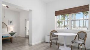 A room with a round table and two chairs, a bed with a bench, a lamp, and a window with a view of palm trees.