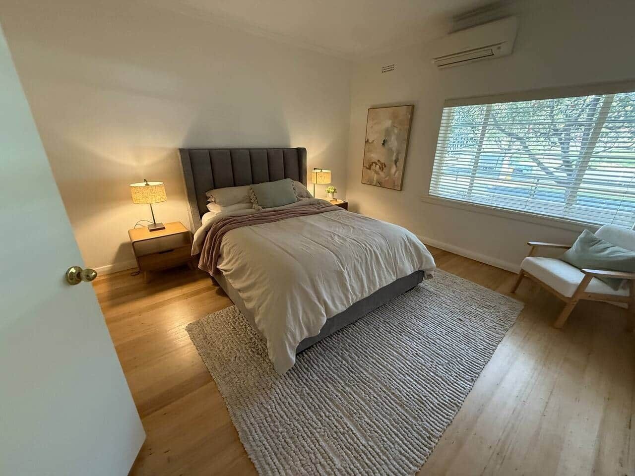 A bedroom with a bed, bedside lamps, a nightstand, a chair, a window with blinds, and a painting on the wall.