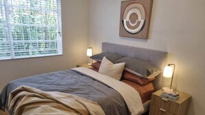 Un dormitorio con cama, mesitas de noche, una obra de arte en la pared y una ventana con persianas.