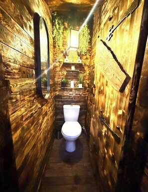 A narrow wooden bathroom with a toilet, shelves with plants, and a mirror. - The Lobster Pot Scarbrough luxury Holiday Let (Scarborough)