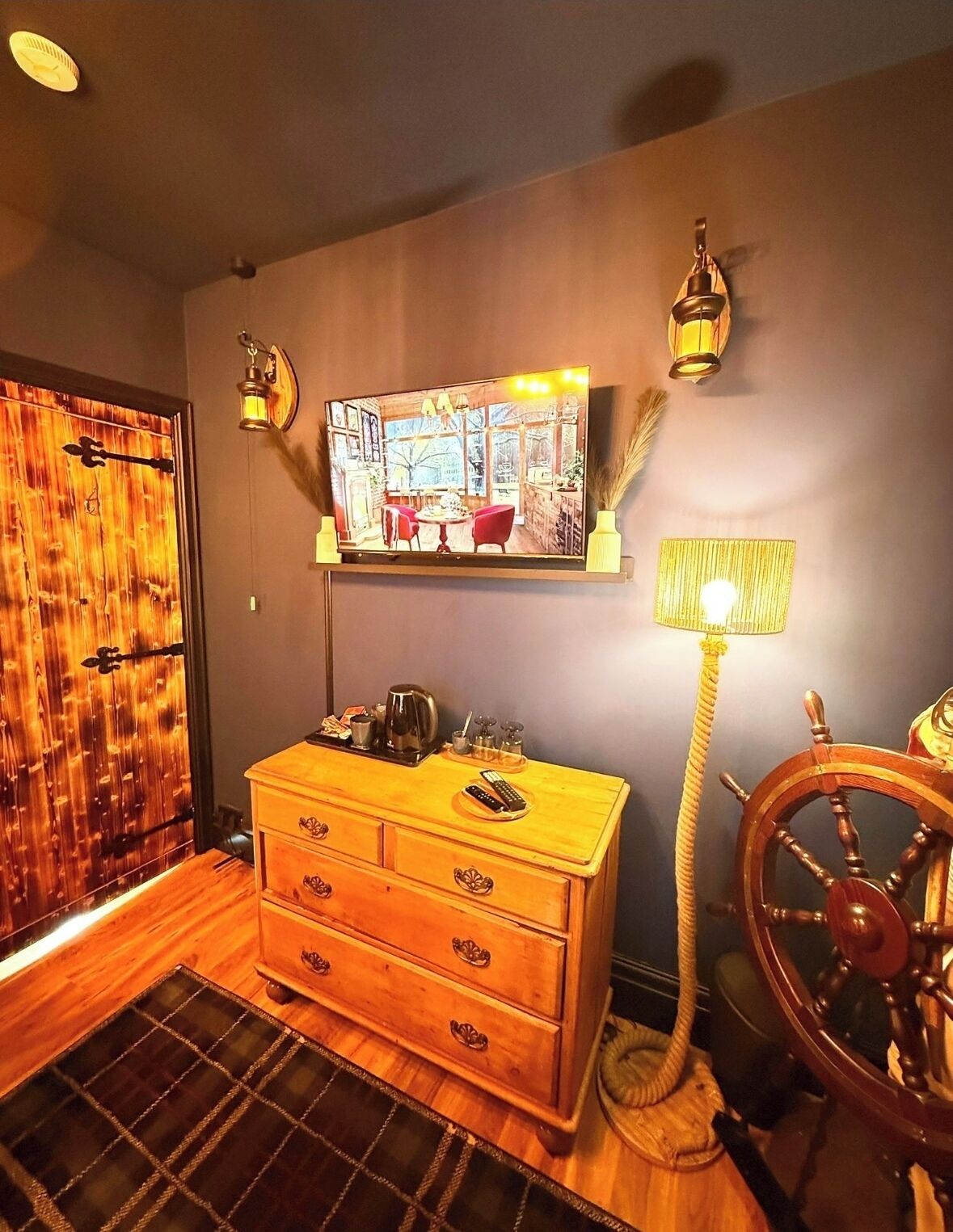 A room with a wooden chest of drawers, a ship's wheel, and a television mounted on the wall displaying a room interior.
