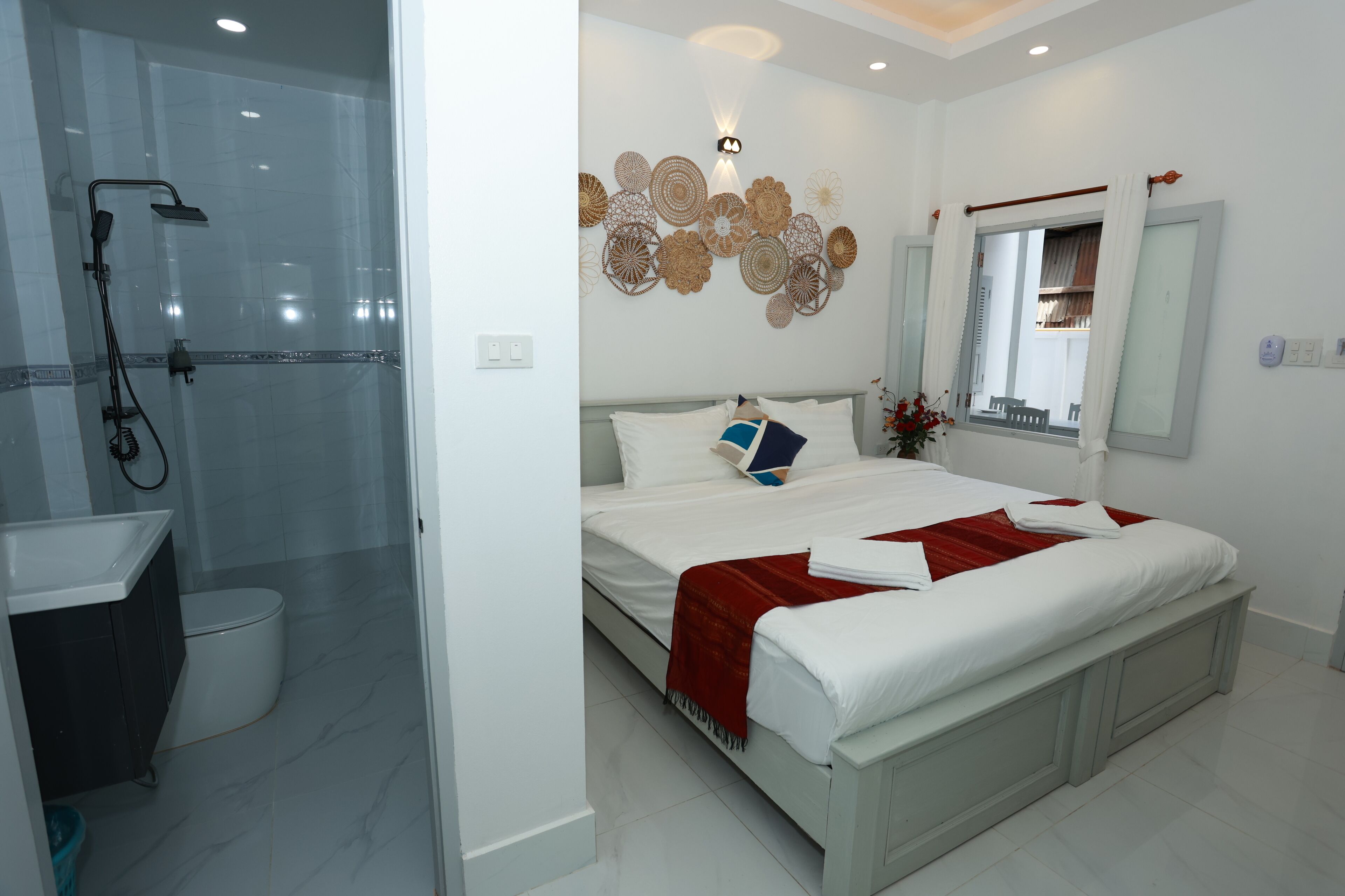 Una habitación de hotel moderna con cabina de ducha de vidrio, un inodoro blanco, una cama bien hecha con una manta roja y un elemento decorativo en la pared sobre la cama.