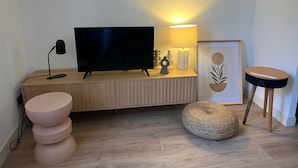 Una sala de estar moderna con un televisor de pantalla plana, una lámpara, una obra de arte enmarcada, un otoman redondito y un taburete pequeño.