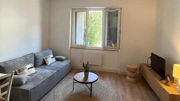 Una sala de estar moderna con un sofá gris, una mesa de centro redonda con una planta, piso de madera, ventilador de techo y una ventana con vista a áreas verdes.