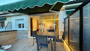 Une terrasse couverte avec une table et des chaises, une porte-fenêtre coulissante donnant sur l’intérieur, et un auvent vert.