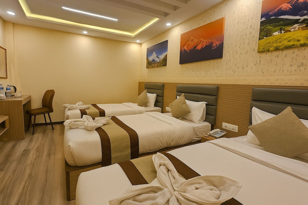A hotel room with two beds, a desk, and a chair. There are paintings on the wall and a ceiling light fixture.