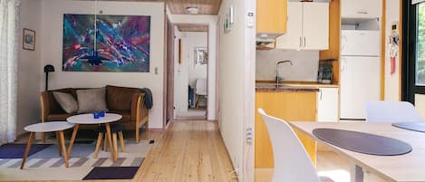 A modern interior with a wooden floor, a dining area with white chairs, a kitchen with white cabinets, and a living area with a brown sofa and a colorful abstract painting.