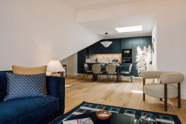 Una sala de estar moderna con un sofá azul, una mesa de comedor con sillas, una cocina al fondo y un tragaluz.