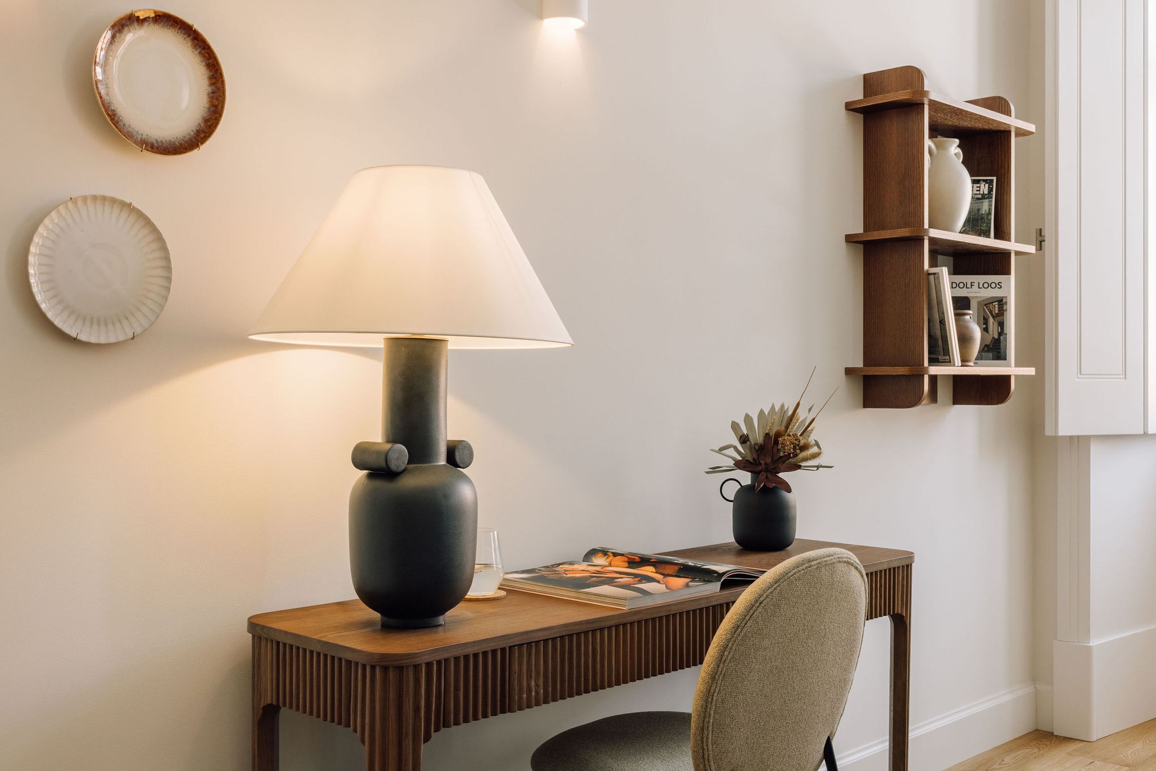 Uma mesa moderna com uma luminária, uma cadeira, uma pequena planta e uma estante em frente a uma parede lisa.