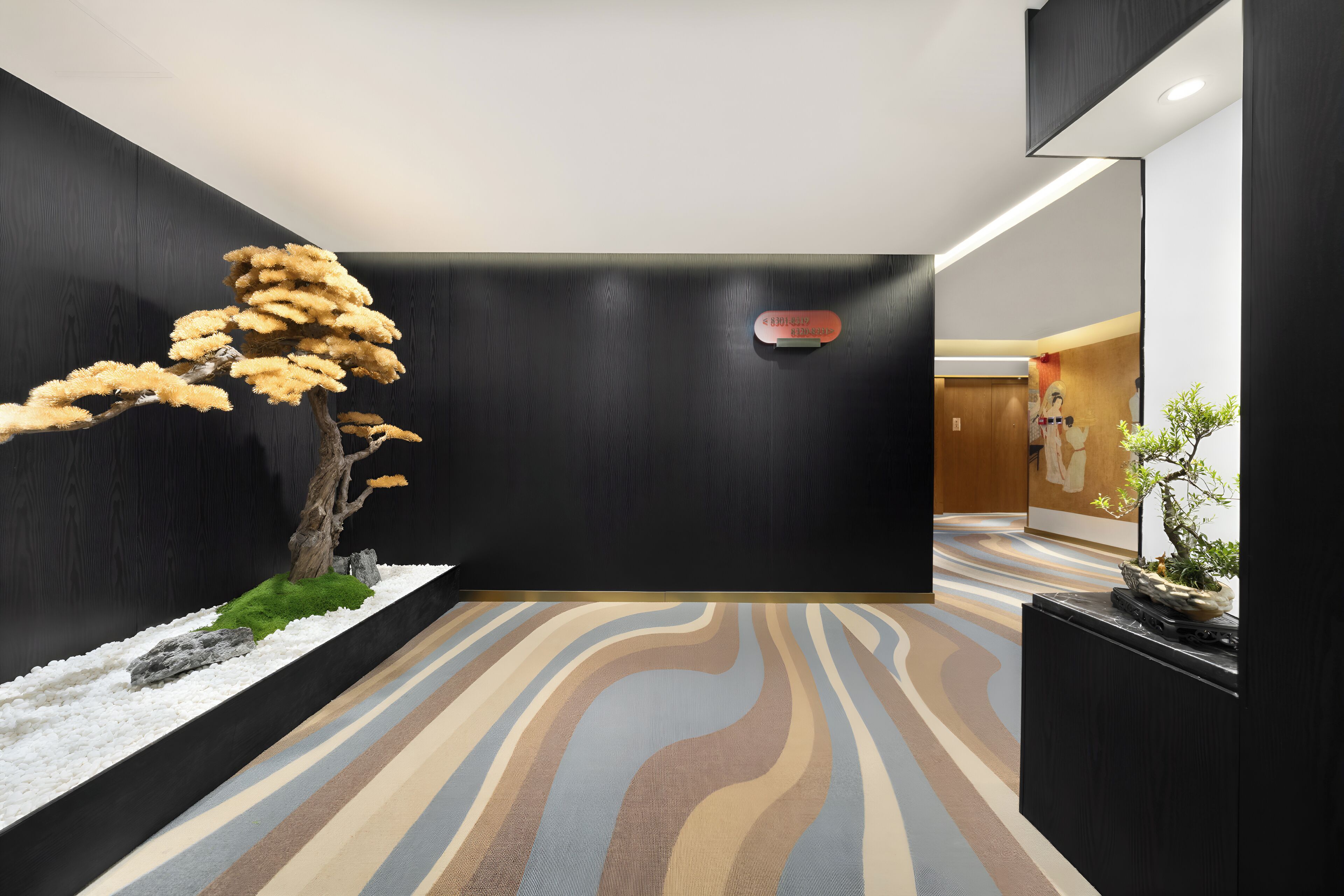 Ein modernes Interieur mit minimalistischem Design, das eine große Baumskulptur, einen gemusterten Teppich und eine dunkle Wand mit einer eingelassenen Deckenleuchte zeigt.