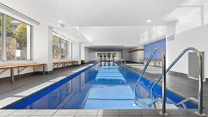 A modern indoor swimming pool with clear glass walls, a long pool, and a seating area with benches. - Central Lakeview Apartments (Canberra)