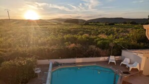 Une piscine rectangulaire bordée d’une terrasse carrelée, entourée de mobilier d’extérieur blanc, et offrant une vue sur un paysage vallonné au coucher du soleil.