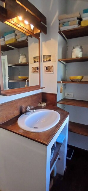 A bathroom with a white sink, wooden vanity, and shelves with towels and toiletries. - Spacious villa with pool and views of the sea and garrigue (Frontignan)